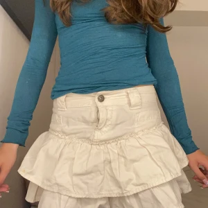 Vit volangkjol med jeansdetaljer - Supersöt vit kortkjol med volanger och jeansinspirerad midja. Kjolen har knapp och dragkedja framtill samt hällor för bälte. Perfekt för dig som vill ha en lekfull och trendig look med extra volym.