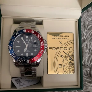 Fredric Tourbillon klocka silver/blå/röd - Snygg mekanisk klocka från Fredric Tourbillions med robust silverfärgad länk i metall. Urtavlan är svart med tydliga index och datumvisning. Ringen runt urtavlan är tvåfärgad i blått och rött, vilket ger en sportig och cool look. Kommer i originalbox med certifikat.