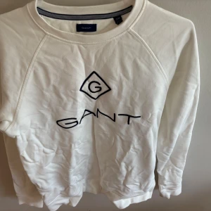 Vit sweatshirt från Gant - Säljer en stilren vit sweatshirt från Gant med broderad logga i svart framtill. Tröjan har rund halsringning och långa ärmar, perfekt för en clean och avslappnad look. Tillverkad i mjuk bomull som känns skön mot huden.