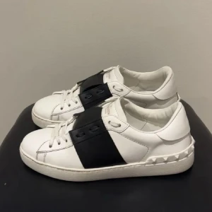 Valentino Open sneakers vit/svart skinn - Snygga Valentino Open sneakers i vitt skinn med bred svart rem över snörningen och ikoniska nitar på hälen. Klassisk rund tå och platt sula. Perfekta för dig som gillar stilrena och lyxiga detaljer.
