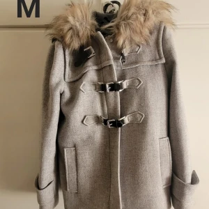 Grå kappa med fuskpäls och spännen - Stilren grå kappa med stor huva och fluffig fuskpäls runt kragen. Jackan har snygga spännen framtill och två fickor. Perfekt för dig som vill ha en klassisk look med en twist. Passar till både jeans och kjol.