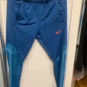 Blå Nike Dri-FIT träningsbyxor M - Snygga blå träningsbyxor från Nike med Dri-FIT-teknologi och slim fit-passform. Byxorna har elastisk midja med snörning, fickor och mönstrade detaljer på benen. Perfekta för träning eller sportiga dagar. Materialet är polyester med lite elastan för extra stretch.