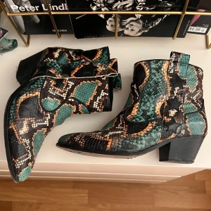 Färgglada cowboy boots med ormmönster - Unika cowboy boots i syntetmaterial med grönt, svart och orange ormmönster. Skorna har spetsig tå, låg klack och svarta stjärndetaljer längs skaftet. 