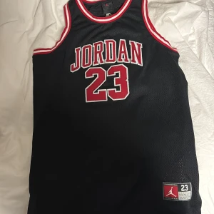 Jordan 23 basketlinne svart/röd - Säljer ett klassiskt Jordan 23 basketlinne i svart med röda och vita detaljer. Stort JORDAN-tryck och nummer 23 fram och bak, samt Jumpman-logga. Ärmlös modell i meshmaterial som andas, perfekt för sport eller streetwear. Samt röda Jordan shorts