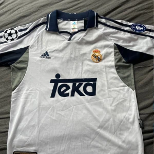 Real Madrid Figo 10 Adidas tröja - Klassisk Real Madrid fotbollströja med Figo 10 på ryggen. Vit med mörkblå detaljer, broderat klubbmärke och Champions League-patchar på ärmarna. Adidas-logga och Teka sponsortryck framtill. Skön och lätt polyester, perfekt för samlare eller fans.