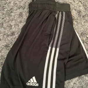 Adidas shorts svart & röd, strl M - Två par Adidas Primegreen shorts i storlek M. Ena paret är svarta med vita ränder längs sidorna och klassisk Adidas-logga, det andra är röda med vita ränder och logga. Båda har elastisk midja och är tillverkade i lätt polyester, perfekta för träning eller fotboll. 100kr/st