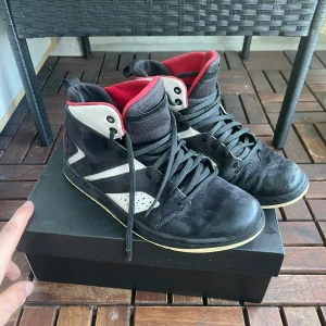 Nike Air Jordan 1 svart/vit/röd - Snygga Nike Air Jordan 1 sneakers i svart mocka med vita detaljer och röd insida. Klassisk high-top modell med svart snörning och den ikoniska Jumpman-loggan broderad bak. Perfekta för dig som gillar streetstyle och vill sticka ut.