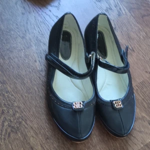 Svarta ballerinaskor  - Stilrena svarta ballerinaskor med rund tå och dekorativt guldfärgat BB-spänne framtill. Skorna har en klassisk rem över foten och är tillverkade i ett slätt material med ljus innersula.