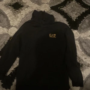 Svart EA7 hoodie med gul logga - Svart hoodie från EA7 med gul logotyp på bröstet. Klassisk huva och känguruficka framtill. Perfekt för dig som gillar stilrena och sportiga plagg. Mjuk och bekväm, passar till många olika outfits.