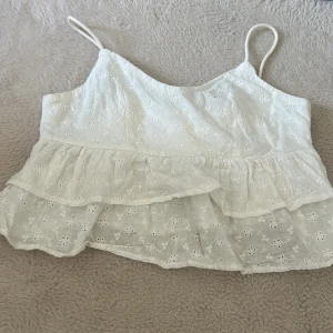 Vit volangig broderad linnetopp - Supersöt vit linnetopp med tunna axelband och broderade blommor. Toppen har dubbla volanger nertill som ger en luftig och romantisk känsla. Perfekt för varma dagar och somriga outfits.
