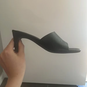 Svarta klackade sandaler från C. Jean. Strl 37 - Svarta sandaler med klack från C. Jean. Snygg minimalistisk design med öppen tå och bred rem över foten. Skorna har fyrkantig tå och är tillverkade i syntetmaterial. Perfekta för dig som gillar stilrena och trendiga skor.