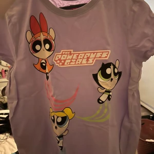 Lila Powerpuff Girls t-shirt Cartoon Network - Lila t-shirt med stort Powerpuff Girls-tryck framtill. Motivet visar Blossom, Bubbles och Buttercup i färgglada toner. Rund halsringning och korta ärmar. Perfekt för dig som älskar retro och tecknade serier.