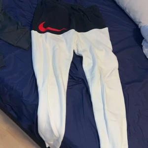 Nike mjukisbyxor svart/vit med röd logo - Snygga mjukisbyxor från Nike med svart överdel och vita ben. Stor röd Nike-logga på höften och dragsko i midjan. Perfekta för chill eller träning, med skön passform och mjukt material.
