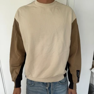 Axel Arigato - Säljer en stilren sweatshirt från Arigato i beige och brunt med svarta detaljer längs ärmarna. Tröjan har rund hals, ribbade muddar och en patch på ena ärmen. Perfekt för dig som gillar en clean och avslappnad look.