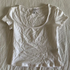 Vit topp från H&M Divided - Enkel och fräsch vit croppad topp från H&M Divided. Modellen har korta ärmar och en djup rundad urringning. Tillverkad i mjuk bomull, perfekt till jeans eller kjol för en clean look.