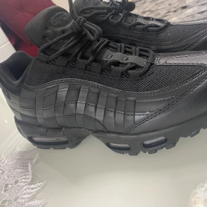 Nike Air Max 95 Triple Black 43 - Nike Air Max 95 i helsvart färg med chunky sula och synliga Air-enheter. Ovandelen är en mix av mesh och skinn med coola vågformade paneler. Klassisk snörning och riktigt streetig vibe. Perfekta för dig som gillar en clean och sportig look.