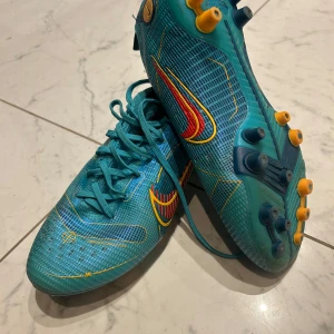 Nike blå fotbollsskor med gula detaljer - Säljer ett par Nike fotbollsskor i blått med gula och röda detaljer. Skorna har snörning, rund tå och platt sula med fasta dobbar i gult och blått. Ovandelen är i syntetmaterial med ett mönstrat rutnät och Nike-logga på sidan. Perfekta för dig som vill sticka ut på planen.
