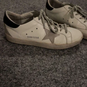 Golden Goose Superstar sneakers vit/grå - Golden Goose Superstar sneakers i vitt skinn med grå mockastjärna på sidan, grå snörning och svart detalj på hälen. Snygg vintage-look med slitna kanter och platt sula. Perfekta för dig som gillar streetstyle och unika detaljer. Kan byta också 