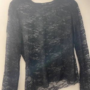 Svart spetsblus med lång ärm - Snygg svart blus i genombruten spets med långärmad modell. Blusen har ett feminint blommönster i spetsen och rund halsringning. Perfekt att styla lager på lager eller med ett linne under.