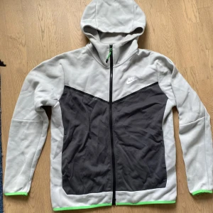 Nike tech fleece hoodie - Nike tech fleece hoodie med dragkedja i grått och svart, med gröna detaljer vid ärmslut och nederkant. Jackan har huva, lång ärm och en sportig look med vit Nike-logga på bröstet. Perfekt för chill dagar eller när du vill ha en casual stil.
