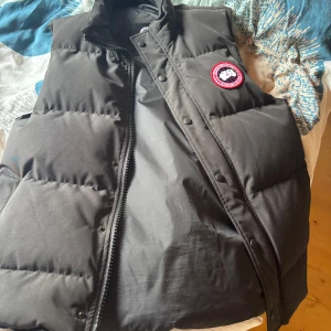 Svart dunväst från Canada Goose - Säljer en svart dunväst från Canada Goose med klassisk logotyp på bröstet. Västen har dragkedja och knappar framtill samt två stora fickor. Perfekt för lager-på-lager under kyliga dagar.