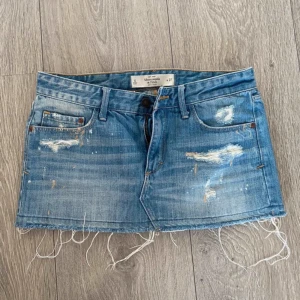 Blå jeanskjol från Abercrombie & Fitch - Säljer en blå jeanskjol från Abercrombie & Fitch med slitna detaljer och rå kant nertill. Kjolen har klassiska fickor fram och bak samt knapp och dragkedja framtill. SKIT SNYGG! tryck inte på köp nu