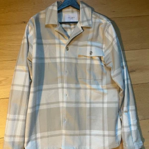 Beige och vit rutig overshirt från Les Deux - Säljer en stilren beige och vit rutig overshirt från Les Deux. Den har klassisk krage, knappstängning framtill och en bröstficka med knapp. Perfekt för dig som gillar en clean och avslappnad look.