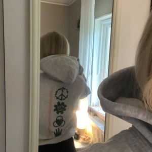 Grå hoodie med grönt tryck - Säljer en grå hoodie med unikt grönt tryck på ryggen och bröstet. Trycket består av olika symboler och orange text. Hoodien har en stor huva och en klassisk känguruficka framtill. Perfekt för en avslappnad stil.