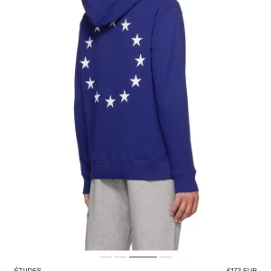 Blå hoodie med stjärnmönster från Études - Säljer en blå hoodie från Études med vita stjärnor i cirkel på ryggen och mindre stjärnmönster på bröstet. Tröjan har huva med dragsko och en stor magficka framtill. Perfekt för dig som gillar streetwear och snygga detaljer.