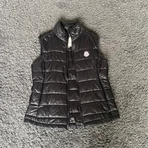 Svart dunväst från Moncler - Säljer en svart dunväst från Moncler med klassisk quiltad design och hög krage. Storlek L dam