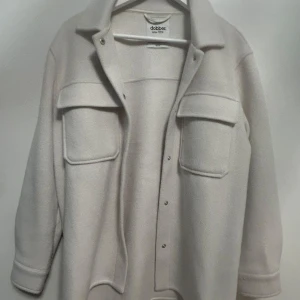 Vit kappa från Dobber - Stilren vit kappa från Dobber med klassisk krage och stora bröstfickor. Jackan har tryckknappar framtill och är långärmad. Perfekt för lager-på-lager och enkel att matcha med olika outfits.