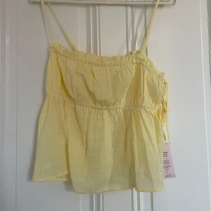 Gul flowy linnetopp från Nelly - Söt ljusgul linnetopp från Nelly med tunna axelband och volangdetaljer. Toppen har knyt i sidorna och en luftig, flowy passform som passar perfekt till sommaren.