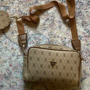 Beige axelväska från Guess - Säljer en beige axelväska från Guess med logomönster och justerbar bred axelrem. Väskan har ett trekantigt Guess-emblem framtill och en liten rund myntväska som hänger på remmen. Fin neutral färg som kan passa till alla outfits .  Säljs pågrund av fick det som present , men inte riktigt min stil. Priset kan diskuteras ! 