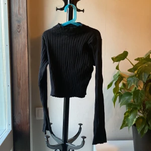 Svart ribbad croppad tröja - Säljer en svart, ribbad och croppad långärmad tröja med tight passform. Perfekt till dig som gillar stilrena basplagg med lite edge. Hög hals och stretchigt material gör den både bekväm och snygg.