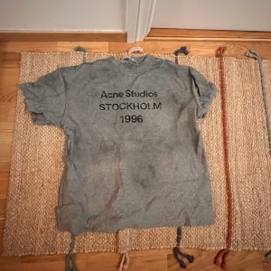 Acne Studios T-shirt - Grå t-shirt från Acne Studios srlk s men sitter som m, med svart tryck på bröstet där det står 'Acne Studios STOCKHOLM 1996'. Klassisk rund halsringning och korta ärmar. Perfekt för en stilren och avslappnad look.