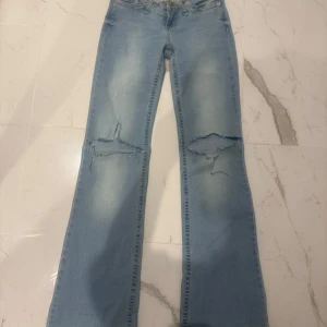 Ljusblå bootcut jeans med slitningar - Säljer ett par ljusblå jeans med bootcut passform och slitna detaljer på knäna. Jeansen har råa kanter nertill och klassiska bakfickor. Perfekta för en avslappnad och trendig look. Har klippt de själva vid knäna !