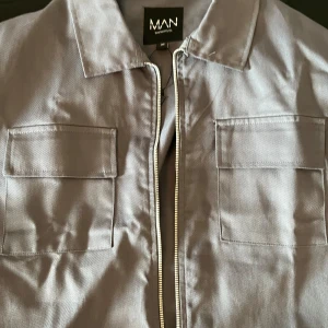 Grå lätt jacka med dragkedja  - Säljer en stilren grå overshirt från MAN med två stora bröstfickor och dragkedja framtill. Jackan har klassisk krage och raka ärmar, perfekt för lager-på-lager-stil.