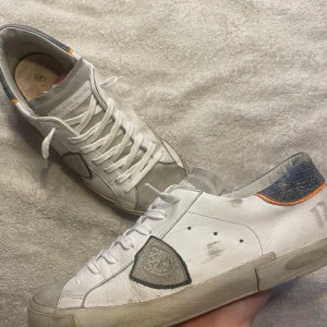 Vita sneakers från Philippe Model - Säljer ett par vita Philippe Model sneakers med grå detaljer och orange inslag på hälen. Skorna har snörning, rund tå och platt sula. Klassisk design med märkets logotyp på sidan och orangefärgad undersida.