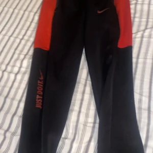 Svarta och röda mjukisbyxor från Nike - Säljer ett par svarta mjukisbyxor från Nike med röda paneler längs sidorna och texten 'Just Do It.' på ena benet. Byxorna har resår i midjan och dragkedjeficka bak. Perfekta för träning eller chill.