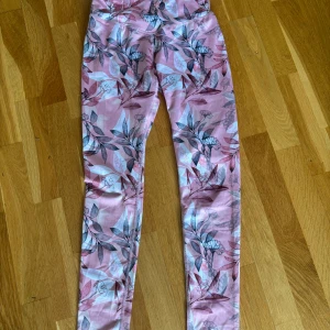 Rosa blommiga träningsbyxor från SOC - Säljer ett par rosa träningsbyxor från SOC med blommigt mönster i vitt, grått och rosa. Byxorna är i stretchigt syntetmaterial, perfekta för gymmet eller löpning. Innerbenslängden och midjemåttet finns med på bilderna, det finns resor i midjan så som mest är midjemåttet 37cm rakt över. Läs bio!💕