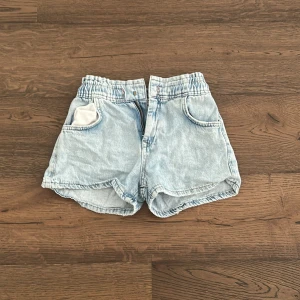Ljusblå jeansshorts från Zara - Ljusblå jeansshorts från Zara med resår i midjan och stora fickor fram. Klassisk femficksmodell i mjukt denimtyg, perfekt för varma dagar. Snygg och avslappnad passform.