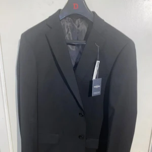 Svart kostym från Dressmann - Säljer en klassisk svart kostym från Dressmann. Inkluderar kavaj, kostymbyxor och skjorta. Nypris 3100kr. Kavajen har två knappar och klassiska slag. Perfekt för bal eller fest. Allt i svart och i stilren design. Vid funderingar eller fler bilder så finns jag alltid i DM. Priset kan diskuteras vid snabbaffär!