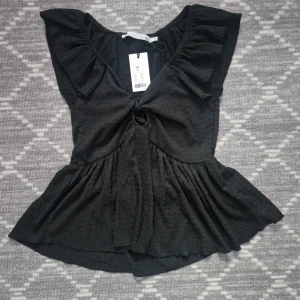 Svart volangtopp med v-ringning - Säljer en svart topp med v-ringning och volangdetaljer vid axlarna. Toppen har en lätt rynkad effekt framtill och är sydd i ett mjukt, lätt material. Perfekt för dig som gillar feminina detaljer och en luftig känsla.