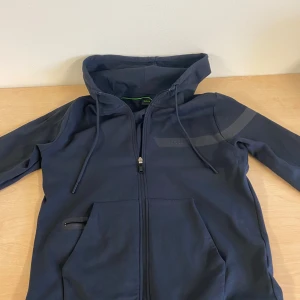 Svart hoodie från BOSS med dragkedja - Säljer en svart hoodie från BOSS med hel dragkedja framtill och huva med dragsko. Tröjan har en diskret logga och dekorativt band på bröstet samt fickor med dragkedja. Perfekt för en stilren och sportig look.