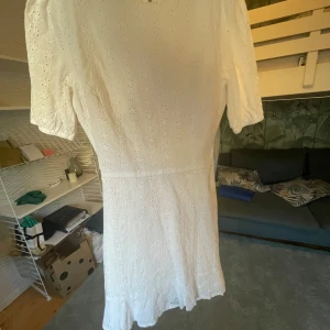 Vit kortklänning med broderade detaljer - Superfin vit kortklänning med broderade blommönster och korta puffärmar. Klänningen har knappar framtill och en öppen rygg med knytband. Perfekt för sommarens alla tillfällen!