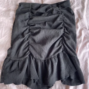 Svart volangkjol från Gina Tricot - Supersnygg svart kjol från Gina Tricot med volangkant och rynkade detaljer framtill. Kjolen har en kort längd och är tillverkad i ett lätt, strukturerat tyg.