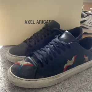 Svarta sneakers från Axel Arigato med broderade fåglar - Svarta sneakers från Axel Arigato med färgglada broderade fåglar på sidorna och tån. Skorna har snörning och vit platt sula. Perfekta för dig som vill ha en stilren men ändå unik look.