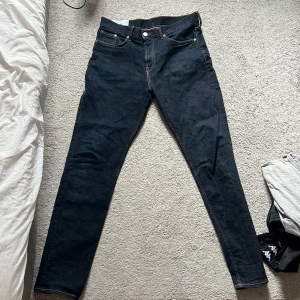 Mörkblå slim fit jeans från H&M - Säljer ett par mörkblå slim fit jeans från H&M. Klassisk femficksmodell med orangea sömmar och raka ben. Jeansen är tillverkade i denim och har en snygg, enkel design som passar till det mesta.