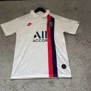 PSG vit fotbollströja från Nike - Säljer en vit Paris Saint-Germain fotbollströja från Nike med blå och röd rand på framsidan. Tröjan har korta ärmar, klassisk krage och PSG-logga på bröstet. Perfekt för dig som älskar fotboll och vill visa ditt stöd för PSG.