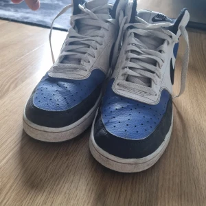 Nike sneakers i blått och svart - Säljer ett par klassiska Nike sneakers med vit bas, blå detaljer och svarta partier. Skorna har snörning, rund tå och platt sula. Perfekta för dig som gillar streetwear och vill ha en stilren look med en färgklick. Dom är lite smutsiga men inte skadad så man behöver bara städa dom